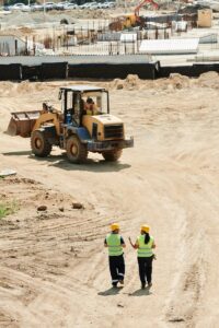 pexels-photo-8961261-8961261 Construction workers in safety gear walking at a busy site with a backhoe in operation.